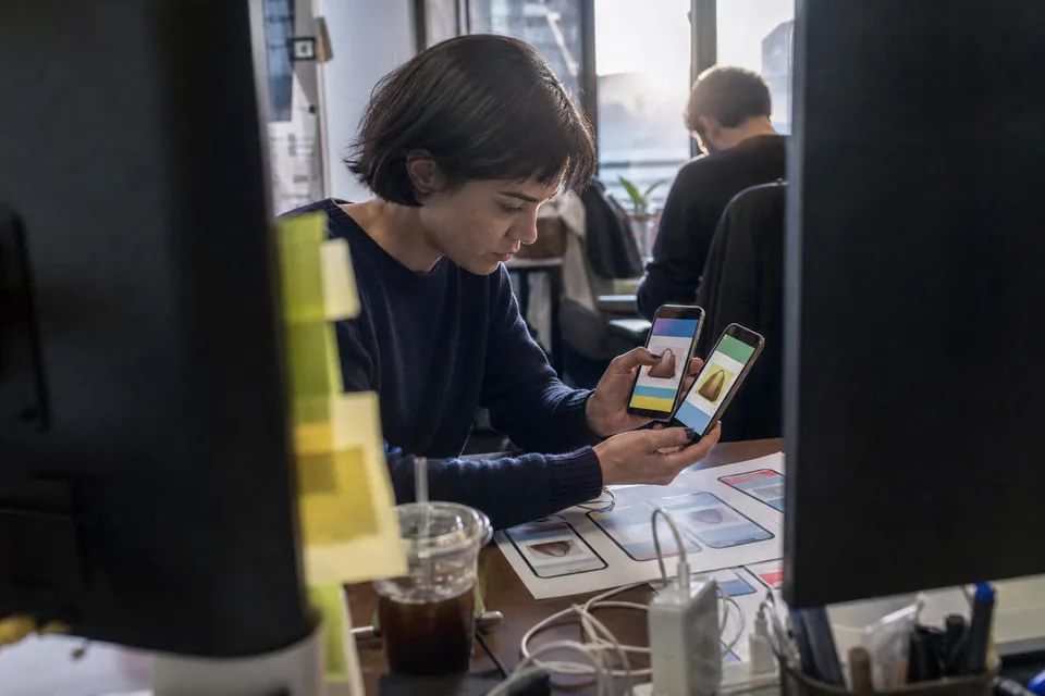 A user acquisition manager at a standing desk compares two phones showing colour-block app screens, monitor out of focus